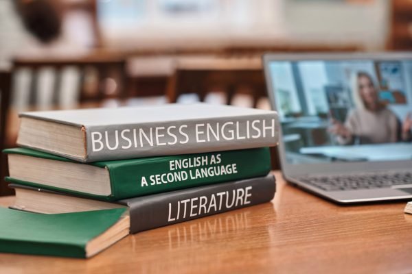 Close up background image of stack of books on English language in school library with video lecture on laptop screen in background, copy space